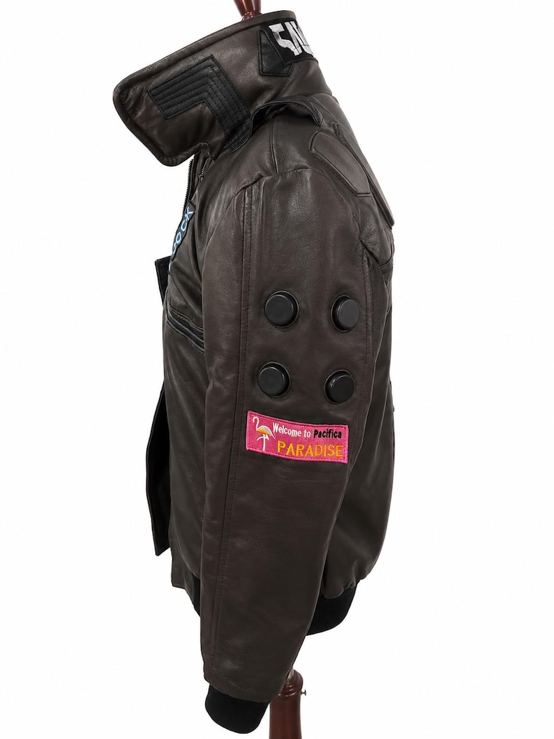 Puede incluir: Una chaqueta de cuero marr&oacute;n oscuro con cuello alto, pu&ntilde;os acanalados negros y cuatro detalles circulares negros en la manga. Un parche rectangular rosa en la parte inferior de la manga muestra un gr&aacute;fico de flamenco y el texto Welcome to Pacifica PARADISE.