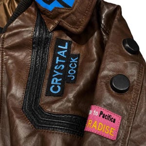 Puede incluir: Una chaqueta de cuero marr&oacute;n con paneles negros texturizados, un detalle de cuello azul y dos botones circulares negros en la manga. Los parches incluyen texto azul CRYSTAL JOCK y un parche rectangular rosa con el texto to Pacifica RADISE.