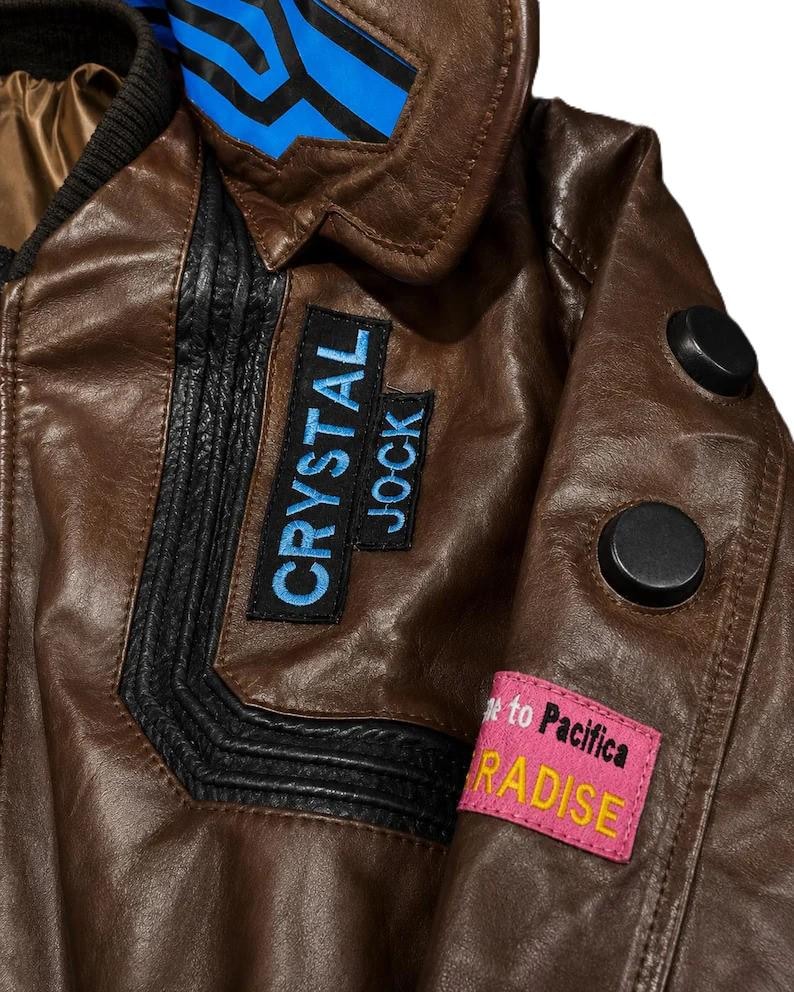 Puede incluir: Una chaqueta de cuero marr&oacute;n con paneles negros texturizados, un detalle de cuello azul y dos botones circulares negros en la manga. Los parches incluyen texto azul CRYSTAL JOCK y un parche rectangular rosa con el texto to Pacifica RADISE.