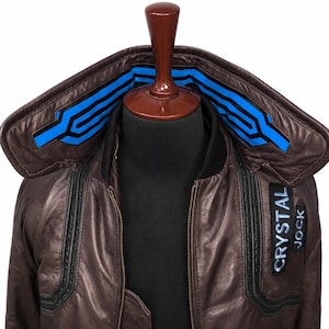 Puede incluir: Una chaqueta de cuero marr&oacute;n con un cuello grande con detalles geom&eacute;tricos en azul brillante y negro. La chaqueta incluye un parche negro en el pecho con el texto blanco CRYSTAL JOCK y un borde acanalado negro a lo largo del cuello y la cremallera.