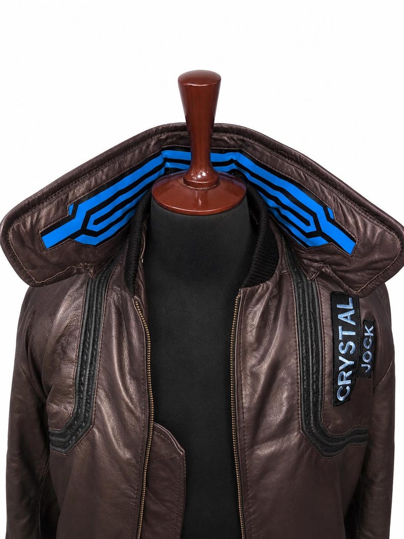 Puede incluir: Una chaqueta de cuero marr&oacute;n con un cuello grande con detalles geom&eacute;tricos en azul brillante y negro. La chaqueta incluye un parche negro en el pecho con el texto blanco CRYSTAL JOCK y un borde acanalado negro a lo largo del cuello y la cremallera.