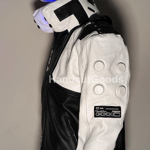May include: A black and white jacket featuring a high collar with black accents, white sleeves with four circular embossed details, and a rectangular black patch on the forearm with white geometric patterns and the text HandoutGoods.
