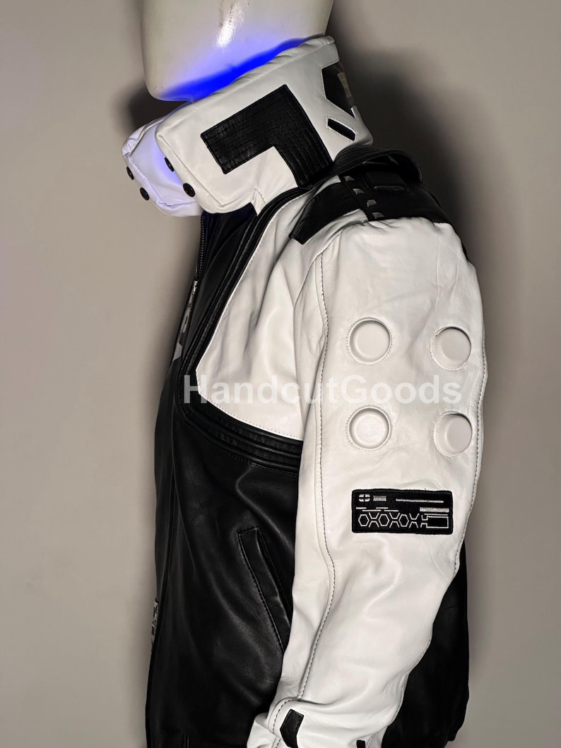 May include: A black and white jacket featuring a high collar with black accents, white sleeves with four circular embossed details, and a rectangular black patch on the forearm with white geometric patterns and the text HandoutGoods.