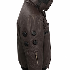 Puede incluir: Una chaqueta de cuero marr&oacute;n oscuro con cuello alto y un parche negro que muestra el texto blanco RAI. La manga incluye cuatro detalles circulares negros y un bolsillo utilitario con cremallera. La chaqueta tiene dobladillo y pu&ntilde;os negros.