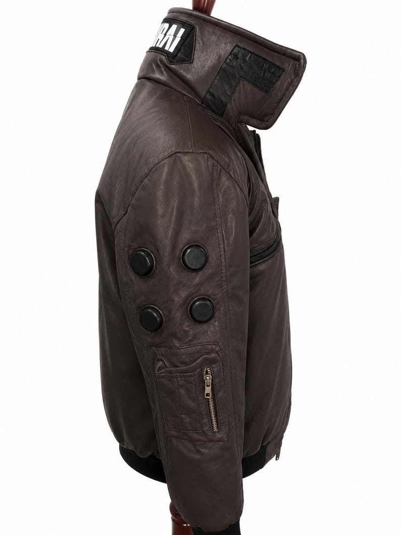 Puede incluir: Una chaqueta de cuero marr&oacute;n oscuro con cuello alto y un parche negro que muestra el texto blanco RAI. La manga incluye cuatro detalles circulares negros y un bolsillo utilitario con cremallera. La chaqueta tiene dobladillo y pu&ntilde;os negros.