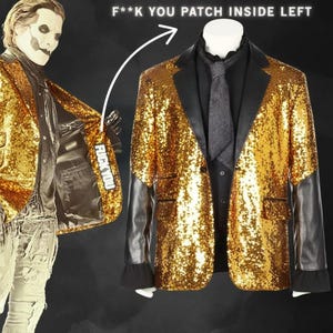 Puede incluir: Una chaqueta dorada con lentejuelas y mangas y solapas de cuero negro. La chaqueta se exhibe en un maniquí con una camisa negra y una corbata gris. El interior izquierdo de la chaqueta tiene un parche que dice "F**K YOU".