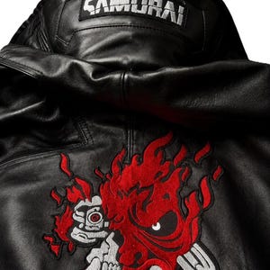 Puede incluir: Una chaqueta de cuero negro con capucha y un parche rectangular en el cuello trasero con el texto SAMURAI en blanco. Debajo del cuello, un gran gr&aacute;fico bordado representa una m&aacute;scara estilizada roja y plateada con llamas y una calavera.