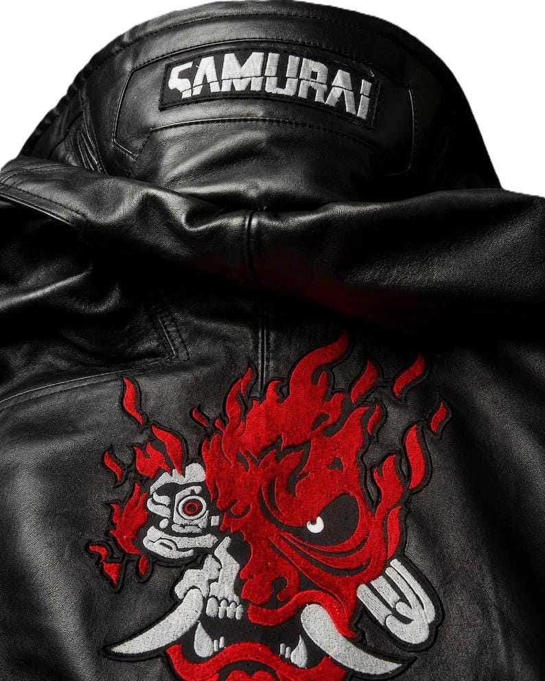 Puede incluir: Una chaqueta de cuero negro con capucha y un parche rectangular en el cuello trasero con el texto SAMURAI en blanco. Debajo del cuello, un gran gr&aacute;fico bordado representa una m&aacute;scara estilizada roja y plateada con llamas y una calavera.