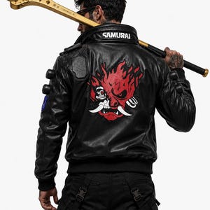 Puede incluir: Una chaqueta de cuero negra con un gran gr&aacute;fico rojo y blanco de una m&aacute;scara oni estilizada en la espalda, con la palabra SAMURAI impresa en el cuello. Una maza dorada con p&uacute;as descansa sobre los hombros, sostenida por una mano tatuada.