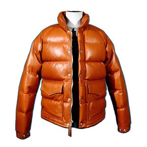 Puede incluir: Chaqueta acolchada de cuero marrón con cuello alto y dos bolsillos delanteros. La chaqueta tiene cierre de cremallera y detalles de botones a presión. La chaqueta se muestra sobre un fondo blanco.