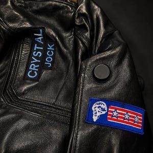 Puede incluir: Una chaqueta de cuero negro con tachuelas piramidales doradas a lo largo de la costura del hombro. Incluye un parche rectangular con el texto bordado en azul CRYSTAL JOCK y un segundo parche rectangular con una calavera blanca y tres estrellas.