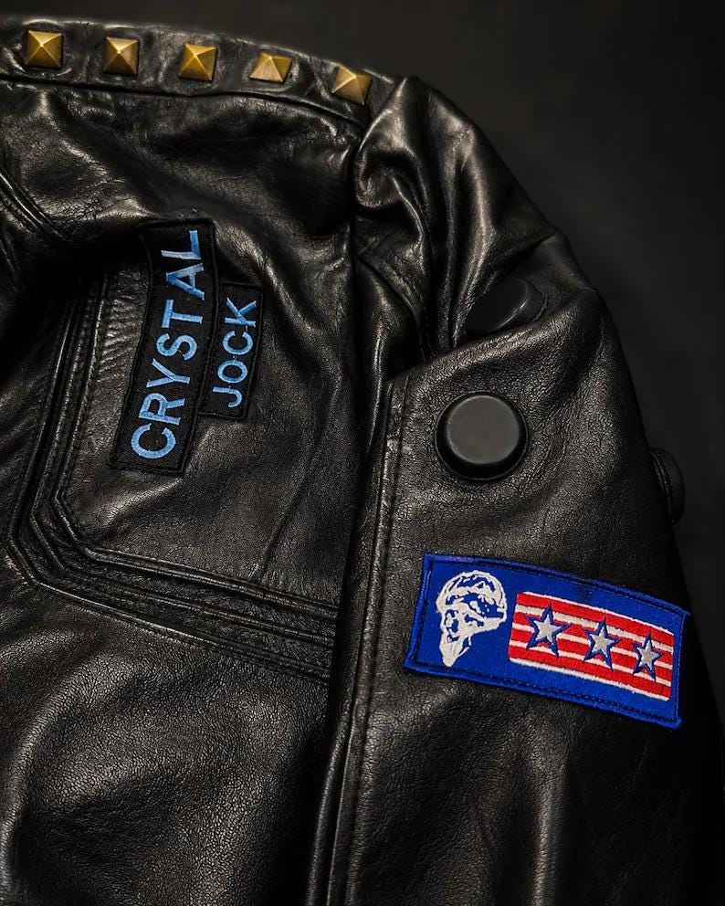 Puede incluir: Una chaqueta de cuero negro con tachuelas piramidales doradas a lo largo de la costura del hombro. Incluye un parche rectangular con el texto bordado en azul CRYSTAL JOCK y un segundo parche rectangular con una calavera blanca y tres estrellas.