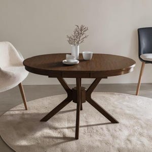 Puede incluir: Una mesa de comedor redonda de madera marrón oscuro con una base central de pedestal y cuatro patas inclinadas. Sobre la mesa hay un jarrón de cerámica blanca con una rama seca, un cuenco pequeño blanco y una taza blanca estriada.