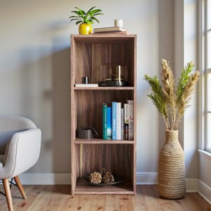 Op de afbeelding: Een houten boekenkast met drie planken en diverse decoratieve items. De bovenste plank bevat een gele pot met een groene plant, boeken en een kaars. De middelste plank heeft een zwart dienblad met gouden accenten. De onderste plank toont een zwart bord met dennenappels.