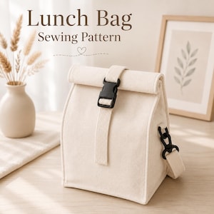 Op de afbeelding: Een crèmekleurige stoffen lunchtas met een oprolbare sluiting die wordt vastgezet met een zwarte plastic gesp. De tas heeft een bijpassende stoffen schouderband bevestigd met zwarte plastic hardware. Tekst bovenaan: Lunch Bag Sewing Pattern.