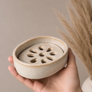 May include: An oval-shaped ceramic soap dish with a speckled cream finish and a removable inner tray featuring a central circular hole surrounded by eight petal-shaped drainage cutouts, creating a functional design for holding soap bars.
