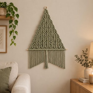May include: A triangular wall hanging made of sage green macrame cord featuring intricate knotted patterns. The piece is suspended from a horizontal wooden dowel and includes a central tassel with long, straight fringe hanging below.