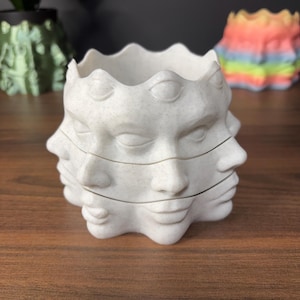 Może przedstawiać: Biała, teksturowana ceramiczna donica z wieloma twarzami. Twarze są ułożone w stos i mają zamknięte oczy i spokojny wyraz twarzy. Obręcz ma falisty wzór. Inne donice są w tle.