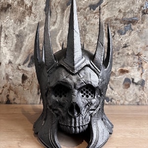 Puede incluir: Un casco negro con forma de calavera con prominentes puntas afiladas. El casco presenta una superficie texturizada detallada y aberturas oculares circulares. El diseño evoca una estética fantástica o gótica.