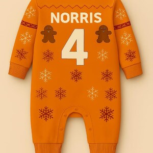 May include: Orange baby jumpsuit with the name "NORRIS" and the number "4" in white. Features gingerbread man and snowflake designs in brown and white. The jumpsuit has long sleeves and snaps at the bottom.