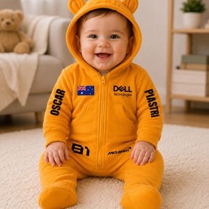 Könnte beinhalten: Ein leuchtend orangefarbener Baby-Strampler mit Kapuze, Bärenohren, Frontreißverschluss und schwarzem Aufdruck OSCAR PIASTRI, 81 sowie McLaren. Auf der Brust befinden sich die australische Flagge und das Dell Technologies Logo.