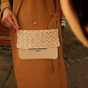 Peut inclure: Un sac à bandoulière en crochet beige avec un fermoir argenté et une bandoulière en chaîne. Le sac a un aspect texturé et tissé et une forme rectangulaire. L'arrière-plan présente un manteau marron.