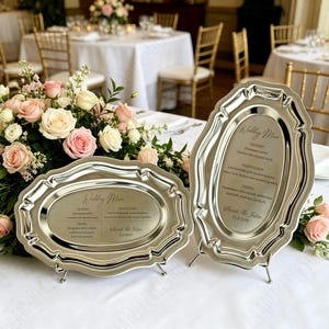 Könnte beinhalten: Zwei silberfarbene Hochzeitsmenü-Tabletts mit verzierten Rändern und kleinen Füßen. Die Tabletts zeigen die Worte "Hochzeitsmenü" und eine Liste von Menüpunkten. Der Hintergrund umfasst ein Blumenarrangement und Tische.