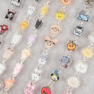 Könnte beinhalten: Eine Sammlung klarer Acryl-Anhänger mit verschiedenen Cartoon-Tierdesigns, darunter Katzen, Hunde und Hasen. Jeder Anhänger ist durch eine silberne Kette verbunden und bildet ein dekoratives Accessoire.