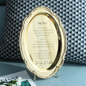 Peut inclure: Un plateau ovale en métal doré avec un bord ornementé et décoratif, servant de présentoir de menu. Le centre présente un texte imprimé en noir listant une entrée, un plat principal et des boissons, se terminant par les noms Preega & Richard.