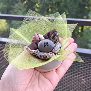 Peut inclure: Ornement floral fait à la main avec une fleur en tissu marron et à carreaux, un bouton gris au centre et un support en résille vert clair. La pièce décorative est tenue dans une main.