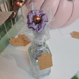 Peut inclure: Petite bouteille en verre transparent ornée d'une fleur violette décorative et d'une perle bronze. La fleur est fixée à la bouteille avec un voile argenté. Une étiquette vierge est suspendue à la bouteille.