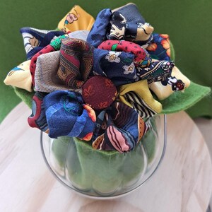 Peut inclure: Un arrangement floral unique fait à partir de cravates recyclées. Le bouquet coloré présente divers motifs et textures, notamment des motifs cachemire et floraux. L'arrangement est présenté dans un vase en verre transparent avec une base en feutre vert, créant une pièce décorative vibrante et écologique.