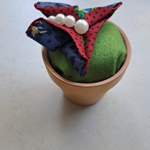 Peut inclure: Un petit pot en terre cuite contient un coussin à épingles en feutre vert, surmonté d'un morceau de tissu décoratif. Le tissu est rouge à pois noirs et accents bleus à motifs, orné de trois perles blanches.