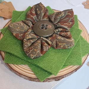 Peut inclure: Une fleur en tissu faite à la main, ornée d'un motif cachemire marron, beige et vert olive, avec un gros bouton au centre. Posée sur des carrés de feutre vert, sur une tranche de bois.