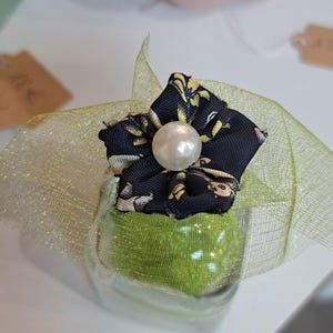 Peut inclure: Un petit pot en verre transparent rempli d'une substance verte, surmonté d'une fleur en tissu noir à motifs floraux et d'une perle. Le pot est enveloppé d'un ruban en résille doré. Une citrouille rose est en arrière-plan.