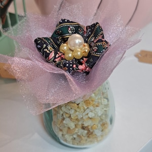 Peut inclure: Composition florale décorative dans un bocal en verre. La fleur est faite de tissu à motifs et de tulle, avec des perles. Le bocal est rempli de petites pierres de couleur claire. Une citrouille rose est en arrière-plan.