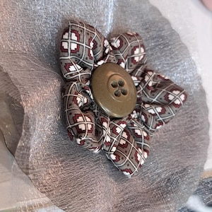 Peut inclure: Une fleur en tissu avec des pétales à motifs gris, bordeaux et blancs, centrée par un gros bouton rond marron. La fleur repose sur une base circulaire argentée et transparente. Un article artisanal.