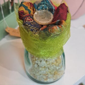 Peut inclure: Un petit bocal en verre rempli de petites pierres de couleur claire. Le dessus du bocal est orné de tissu coloré et d'un filet vert clair, et d'un objet rond et transparent.