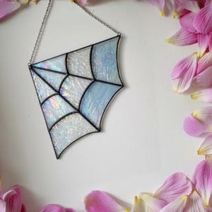 May include: A stained glass suncatcher shaped like a spider web, featuring iridescent white and pale blue glass panels held together by dark metal solder lines. The piece hangs from a thin silver-coloured metal chain attached to the top corner.