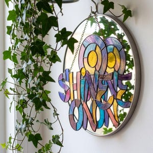 May include: A circular wall mirror featuring a hanging stained glass ornament with the text SHINE ON. The glass piece is composed of multicolored segments in shades of yellow, blue, and purple, suspended by a metal chain from a hook above the mirror.