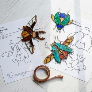 Pode incluir: Padrões de insetos em vitrais e insetos em vitrais acabados em tons de castanho, azul, verde e amarelo. Os padrões são impressos em papel branco com o texto "Flying Bugs Bundle". Há também um rolo de fita de cobre.