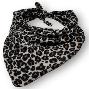 Peut inclure: Un bandana à imprimé léopard dans les tons de noir, marron et crème. Le tissu est noué en haut. Le bandana est de forme triangulaire et est probablement fait d'une matière douce.