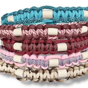 Könnte beinhalten: Fünf bunte Paracord-Armbänder mit weißen Perlen. Die Armbänder sind übereinander gestapelt. Das oberste Armband ist türkis, das zweite ist rosa und burgunderrot, das dritte ist burgunderrot, das vierte ist rosa und lavendelfarben, und das unterste Armband ist beige und braun.