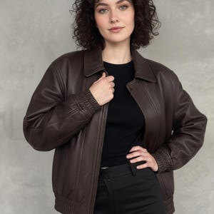 May include: A dark brown leather jacket with a zip fastening, a collared neckline, and ribbed cuffs. The jacket is worn over a black top and paired with black trousers. The overall style is casual and fashionable.