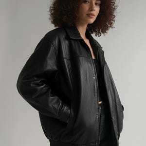 Op de afbeelding: Zwarte leren jas met een kraag, knoopsluiting en zijzakken. De jas heeft een bomber-stijl ontwerp met een geribbelde zoom en manchetten. Het model draagt de jas.