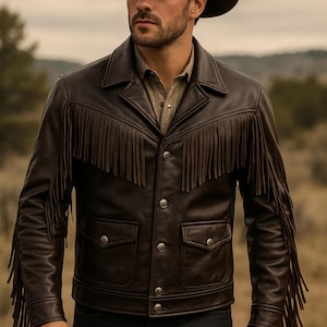 Puede incluir: Una chaqueta de cuero marrón oscuro con flecos en el pecho, los hombros y las mangas. La chaqueta tiene dos bolsillos delanteros y botones plateados. La persona de la imagen lleva un sombrero de vaquero marrón y una camisa abotonada.
