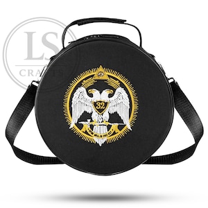 Puede incluir: Bolso redondo negro con asa y bandolera. El bolso presenta un emblema dorado y blanco de un águila bicéfala con el número 32. El emblema está rodeado por un diseño de rayos de sol dorados.