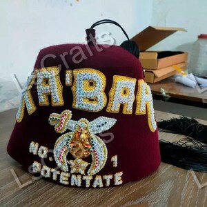 Könnte beinhalten: Ein roter Fez mit goldener Stickerei und Strasssteinen. Der Hut trägt die Aufschrift "K'BAR" und "POTENTATE". Der Hut ist mit einem goldenen und mit Edelsteinen besetzten Emblem verziert.