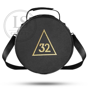 Puede incluir: Bolso redondo negro con un triángulo bordado en oro y el número 32. El bolso tiene un asa superior y una correa para el hombro negra. Fabricado con un material texturizado.