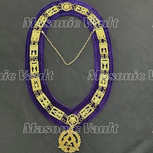 Puede incluir: Un collar masónico dorado con un respaldo de terciopelo morado. El collar presenta intrincados diseños geométricos y símbolos, incluyendo estrellas y cuadrados. Un colgante dorado con la letra "A" cuelga de la parte inferior.